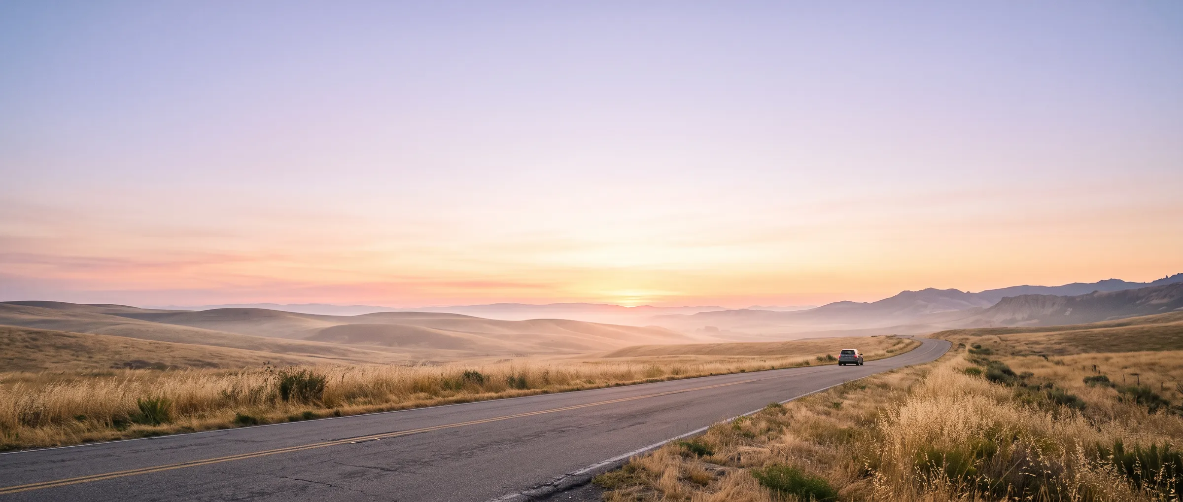An empty road winding through hills toward a lavender-and-peach sunrise, a small car in the distance